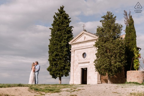 An engaged couple stands embraced in front of the small Chapel of Vitaleta in Tuscany, surrounded by iconic cypress trees and the still, romantic light of the Italian countryside.