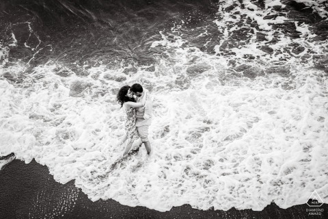 Two lovers are embraced by the crashing waves in Positano, Italy, lost in each other during a candid engagement portrait that captures the raw and pure connection of their romantic bond.