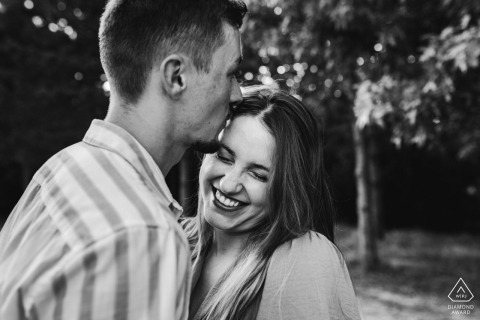 At Lac Vert de Canéjan in Gironde, France, the fiancé affectionately kisses his future wife on the temple, making her laugh during their serene and natural outdoor engagement portrait session.