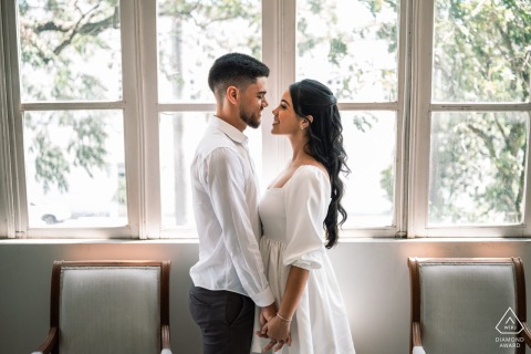 Holding hands in front of a large, grand window at Palácio Deodoro in Alagoas, the couple gazes into each other's eyes, illuminated by the soft light filling the historic room.