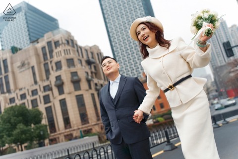 She beams with happiness and lifts her bouquet high as the couple strolls hand in hand through the vibrant city streets along the Bund in Shanghai, China, for their engagement.
