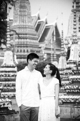 A couple stands close for a portrait in Bangkok, Thailand, their expressions reflecting a deep connection as the city atmosphere subtly frames the background, highlighting their shared happiness.