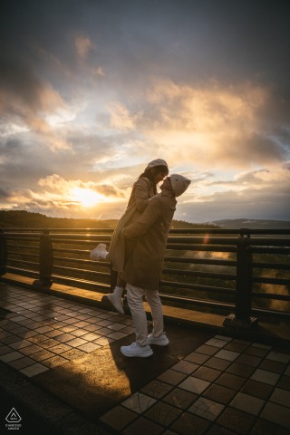 Standing on a bridge in Aomori, Japan, the couple poses at sunset, the fading orange hues creating a gentle backdrop as they hold each other, surrounded by the quiet beauty of the evening.