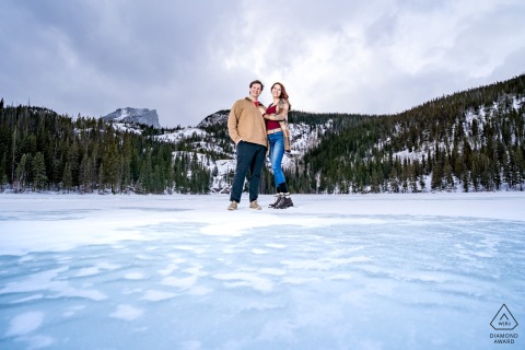 On frozen Bear Lake in Rocky Mountain National Park, Colorado, the couple stands hand in hand surrounded by snowy scenery, the icy expanse and mountains creating a stunning winter landscape for their engagement portrait.