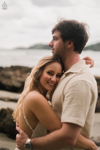 On The Sandy Shoreline In Guarujá, São Paulo, The Couple Enjoys A Relaxed Portrait By The Sea On the sandy shoreline in Guarujá, São Paulo, the couple poses for a relaxed engagement portrait, with soft light and the warmth of their relationship by the ocean.