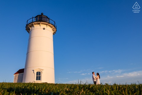 Nestled in grass below Nobska Lighthouse in Falmouth, Massachusetts, the couple stands at the base of the historic beacon, a low angle highlighting their connection amid an expansive late-day coastal sky.