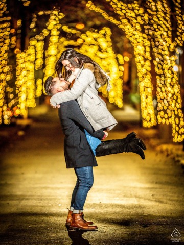 Lifted in his arms, she smiles with joy as the warm glow of Addison Street lights shimmer behind them in Philadelphia, their engagement portrait radiating love and happiness on this evening.
