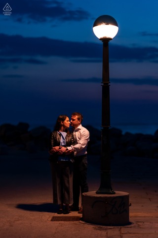 Scene Setter Of A Couple Overlooking The Adriatic Sea During Evening Glow In Piran Slovenia Soft evening glow from a street light settles over Piran, Slovenia, as a couple stands close together for an engagement portrait overlooking the historic coastal town and the calm Adriatic Sea.