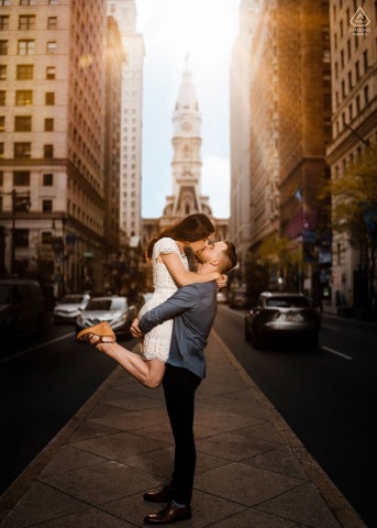 A couple shares a sunset kiss outside Philadelphia City Hall, warm evening light glowing between the surrounding streets and dramatic architecture during their romantic urban engagement portrait.