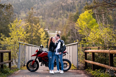 Leaning casually beside their motorbike in Tarvisio, Udine, Italy, a couple shares a relaxed engagement portrait that reflects their adventurous spirit against a scenic alpine town backdrop.