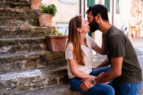 A couple shares a kiss along the waterfront in Grado, Gorizia, Italy, gentle seaside light illuminating their engagement portrait as they sit together in the relaxed coastal atmosphere.