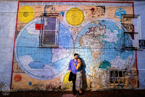 A couple embraces beside a vibrant city wall featuring a colorful painted map at Iron Hill Brewery in Philadelphia, their engagement portrait framed by bold artwork and lively urban textures.