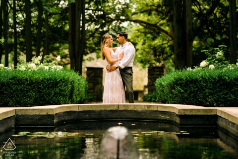 A couple embraces beside a quiet garden fountain at Hunting Hill Mansion in Pennsylvania, their engagement portrait highlighting the lush greenery and peaceful setting surrounding them.