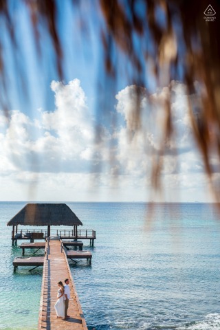 A bride stands at the end of a wooden pier at Viceroy Playa del Carmen, framed by soft foreground leaves, the turquoise water and bright blue sky completing the serene backdrop of her engagement portrait.