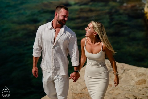 A couple walks hand in hand along a rocky coastline in Cassis, France, their smiles and the vibrant turquoise water of the Mediterranean providing a lively backdrop to their engagement portrait.
