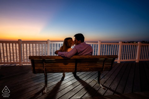 On a pier in Philadelphia, Pennsylvania, a couple kisses at sunset, captured in a backlit engagement portrait with warm light and open space surrounding them.