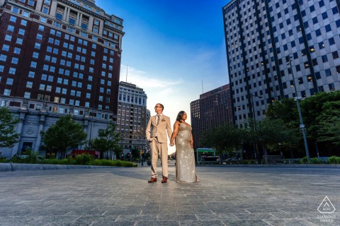 Between city streets in Philadelphia, Pennsylvania, a couple stands hand in hand, shown in an urban engagement portrait with buildings and open sky forming a horz composition.