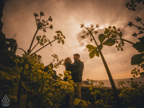 At Portovenere, Liguria, Italy, a couple stands surrounded by flowers, presented in a natural light engagement portrait that uses floral details as a central visual element.