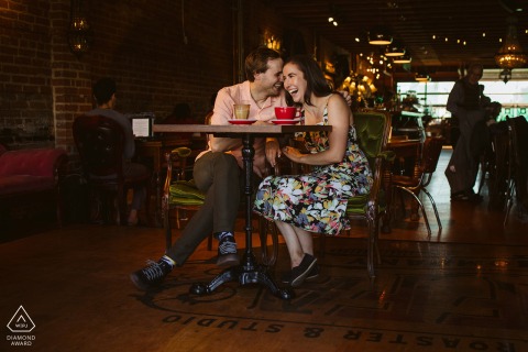 At a coffee shop in central Edmonton, Alberta, a couple shares a laugh with their heads close together at a table, creating a warm and intimate engagement portrait.