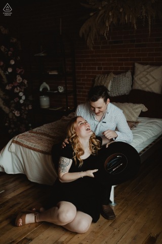 In a downtown Edmonton studio, a couple sits at the base of a bed, playfully interacting with a cowboy hat in a candid, relaxed engagement portrait.