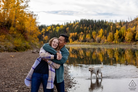River Valley, Edmonton, Alberta – A man hugs his partner from behind as their dog plays in the river nearby, capturing a playful, intimate engagement shot.
