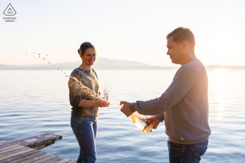 Standing along the serene shores of the Lake of Zoug in Switzerland, the happy couple celebrates their upcoming wedding by popping a bottle of champagne against the water.