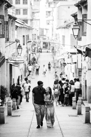 In Macau, the couple walks up a narrow business street in black and white, with tall vertical lines emphasizing a stylish, cinematic portrait and elegant city setting.