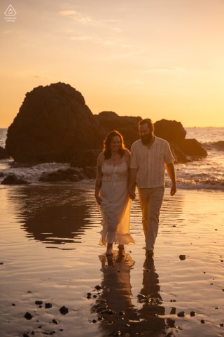 In Costa Rica, the couple walks in front of the ocean at sunset, their silhouettes framed by warm light, calm water, and a romantic coastal backdrop.