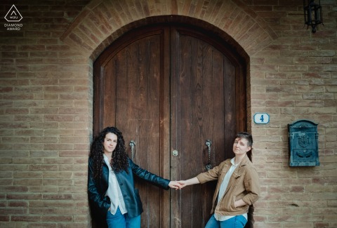 In Colle Val D Elsa Tuscany The Couple Holds Hands Beneath A Brick Archway For Engagement Session In Colle Val d'Elsa, Tuscany, the couple holds hands beneath a brick arch and wooden door, creating an elegant engagement portrait with historic textures and warm charm.