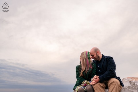 In Trieste, Italy, the couple cuddles while sitting on rocks, sharing a tender coastal portrait with natural textures, close affection, and a quiet seaside feel.