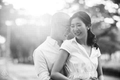 Black and white pre-wedding engagement portrait of a smiling couple embracing beneath soft natural light in Bangkok, Thailand.