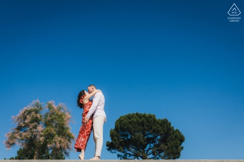 A couple embraces atop a stone wall beneath a cloudless blue sky during their engagement portrait session in Cap Ferret, Gironde, France.