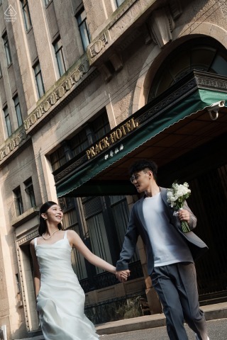 The Couple Walk Past The Peace Hotel At The Bund In Shanghai, China An engaged couple walks past the historic Peace Hotel while holding hands during a stylish pre-wedding session at the Bund in Shanghai, China.