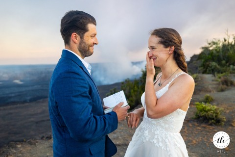 The bride wipes away a tear yet beams with joy as she shares heartfelt vows with the groom on the stunning rim of Hawaii Volcanoes National Park in Hawaii, USA.