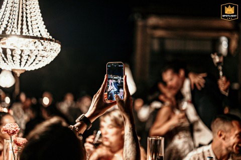 At Podere Palazzo courtyard, Cesena, Italy, the spouses kiss, captured through a guest's phone screen, with chandeliers illuminating the courtyard.