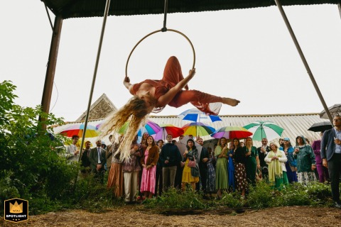 At a family farm in Bridport, Dorset, England, the bride's sister, an aerial artist, performs on a hoop in a chicken shed as guests watch with colorful umbrellas in the rain.