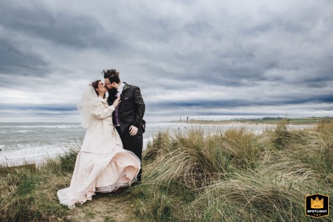 Bride and groom share a romantic kiss on Alnmouth Beach, Northumberland, UK, with the chilly North Sea behind.