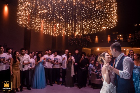 At Espaço Welucci – São Paulo, Brasil, the bride and groom share their first dance, positioned to the far right, with a row of guests filling the background.