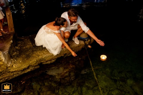 At a restaurant by the sea in Symi, a bride and groom crouch, releasing a floating, burning candle into the water.