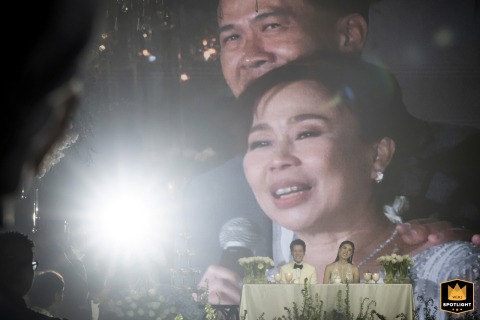 At Four Seasons Bangkok, a couple is seated at the head table, with a massive projection screen behind them showing an emotional woman speaking into a microphone.