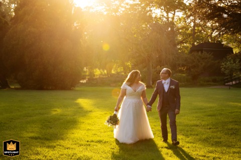 At Fanhams Hall, during golden hour, the bride and groom walk hand-in-hand on the grass by trees, sun setting behind them.
