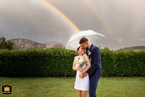In Varces city, Isère, France, a laughing bride and groom stand under a rainbow, holding a white umbrella.