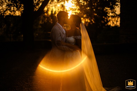 At Castello di Mugnana, Tuscany, Italy, a kissing bride and groom are backlit by a golden, copper-toned sunset with flares.