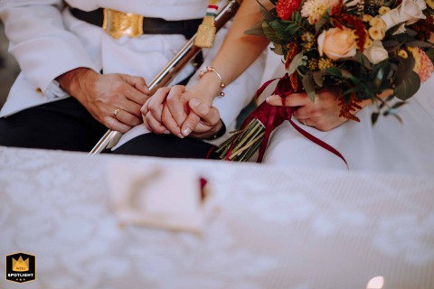 At their Budapest ceremony, a tender close-up captures the bride and groom holding hands tightly—her bouquet resting on her lap and him in ceremonial uniform—highlighting a powerful, quiet moment of unity and love.
