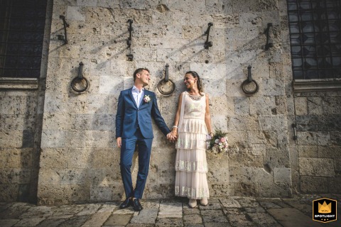 At Piazza del Campo in Siena, Tuscany, the couple is captured in a romantic portrait beside an old stone-carved wall, accented with iron rings mounted above, blending timeless charm with the city’s historic elegance.