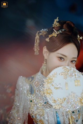 At Jing'an Shangri-La in Shanghai, China, the bride, dressed in traditional Chinese wedding attire and holding an ornate fan, exudes timeless elegance. A softly blurred background, created by red glass, enhances the refined beauty of this portrait.