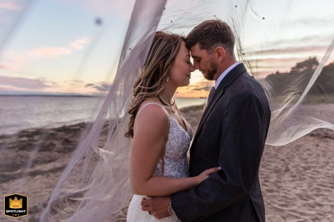At The Whaf in Falmouth, an intimate couple poses on the beach at sunset, beautifully framed by soft golden light and the delicate movement of the bride’s veil.