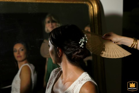 At Domaine de Lalanne in Occitanie, France, bridal preparations unfold in a serene setting. Reflected in surrounding mirrors, another person gently holds a hand fan for the bride, capturing quiet care, elegance, and anticipation.