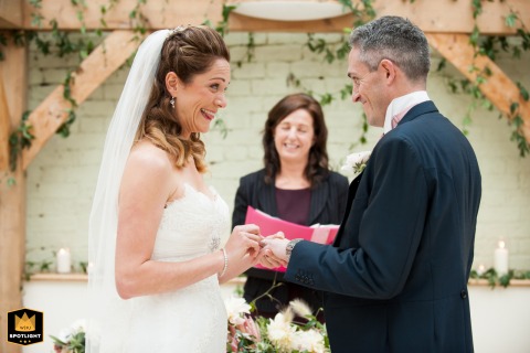 At Gaynes Barn in Essex, the bride playfully sticks out her tongue while trying to place the wedding ring on the groom's finger. The candid shot captures a moment of lightheartedness.