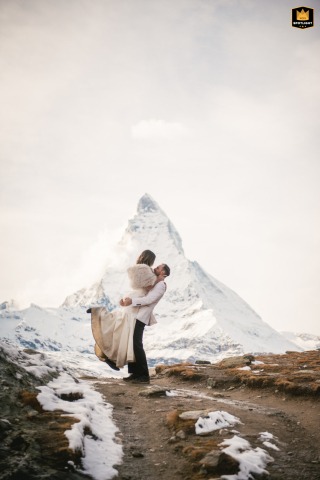 A couple kisses in Zermatt, Switzerland, with the iconic Matterhorn visible in the snowy background. The romantic moment captures their affection amidst the stunning winter landscape.